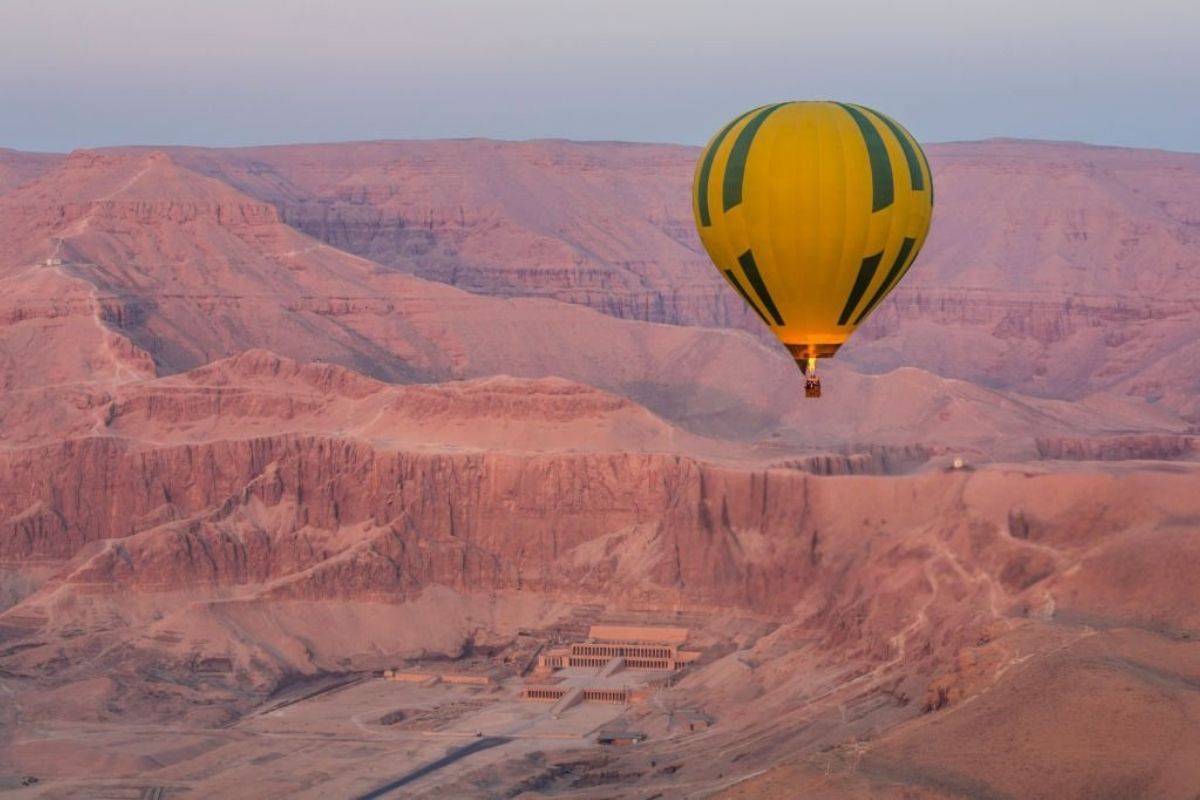 Yellow and green hot air balloon floating over the desert cliffs of Luxor with Hatshepsut Temple below