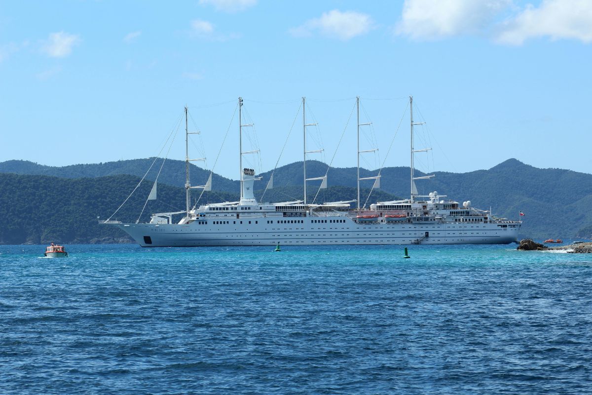 Large luxury cruise ship with white sails docked in clear blue waters with lush green mountains in the background